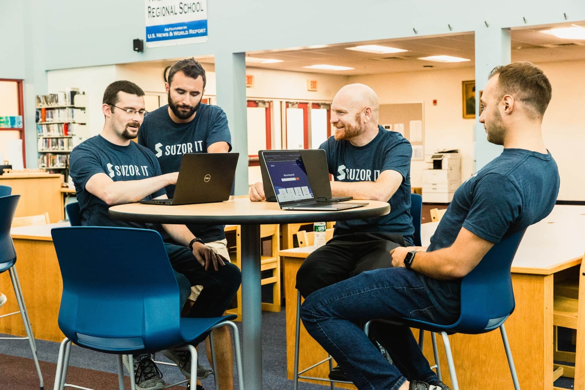 Four Suzor IT technicians in navy Suzor tees working together on laptops at a table in a school library